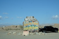 A colorful handmade sign reading 'Salvation Mountain' and 'God Never Fails' stands in a desert landscape. The sign is adorned with bright painted flowers and sits atop a mound of stones. A black pickup truck is parked nearby under the clear blue sky.