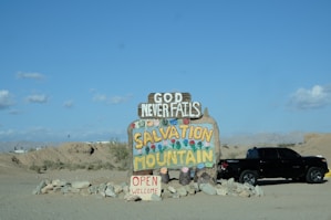 A colorful handmade sign reading 'Salvation Mountain' and 'God Never Fails' stands in a desert landscape. The sign is adorned with bright painted flowers and sits atop a mound of stones. A black pickup truck is parked nearby under the clear blue sky.