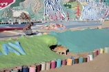 A person in a plaid shirt is crouched down on a colorful, painted surface, possibly working on a mural or art project. The background is filled with vibrant abstract patterns and nature motifs, including trees and flowers, rendered in bright colors. Numerous paint cans, both used and unused, are lined up along the ground in front of a painted bank, where a cat is emerging from a small tunnel.