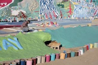 A person in a plaid shirt is crouched down on a colorful, painted surface, possibly working on a mural or art project. The background is filled with vibrant abstract patterns and nature motifs, including trees and flowers, rendered in bright colors. Numerous paint cans, both used and unused, are lined up along the ground in front of a painted bank, where a cat is emerging from a small tunnel.