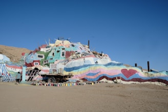 A colorful, painted structure resembling a small hill or mountain with various messages of love and spirituality. The surface is adorned with colorful stripes, hearts, and phrases such as 'God is Love' and 'Love is Universal.' The sky is clear, indicating daylight. A person is walking near the structure, further emphasizing its large and artistic nature.