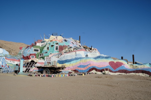 A colorful, painted structure resembling a small hill or mountain with various messages of love and spirituality. The surface is adorned with colorful stripes, hearts, and phrases such as 'God is Love' and 'Love is Universal.' The sky is clear, indicating daylight. A person is walking near the structure, further emphasizing its large and artistic nature.
