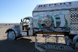 An old truck is covered with colorful religious and spiritual messages and drawings. The vehicle is painted predominantly white with various murals and texts, including phrases like 'God is Love' and 'Repent.' Decorations include hearts and biblical references. The setting is a barren desert landscape, and there is a worn sign saying 'Please Keep Off.' The truck’s interior is visible with its open door.