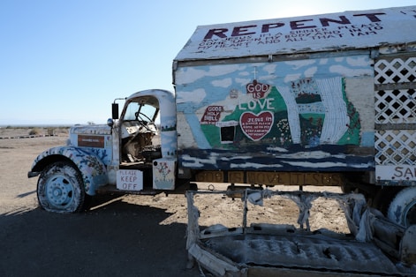 An old truck is covered with colorful religious and spiritual messages and drawings. The vehicle is painted predominantly white with various murals and texts, including phrases like 'God is Love' and 'Repent.' Decorations include hearts and biblical references. The setting is a barren desert landscape, and there is a worn sign saying 'Please Keep Off.' The truck’s interior is visible with its open door.