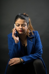 A woman with a serious expression is holding a smartphone to her ear, dressed in a blue blazer with a black top. She appears to be deep in thought during a phone call. The background is dark, enhancing the focus on her expression and attire.