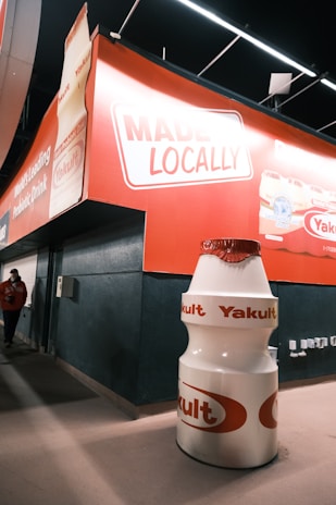 A large Yakult bottle display stands prominently in front of a wall featuring bold red signage with the text 'MADE LOCALLY.' The area appears to be part of an indoor setting with overhead lighting. A person wearing a red jacket is walking nearby, partially visible on the left side.