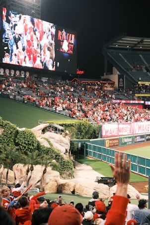 A crowded stadium filled with people wearing predominantly red clothing. A large screen displays images of the crowd along with the text 'Rally Time.' The surrounding area includes a section with rocks and greenery, adding a natural element to the environment.