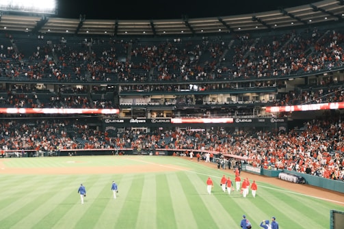 A baseball stadium filled with spectators, mostly concentrated in the stands. The outfield grass is well-maintained and vividly green. Several players wearing red uniforms are walking on the field, along with a couple in blue. Advertisements are visible around the stadium.