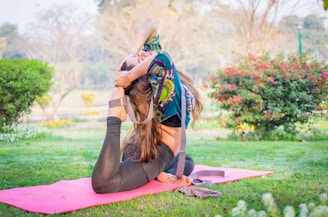 A woman is performing a yoga pose on a pink mat in a vibrant green garden. The pose involves bending backwards, with one leg stretched behind her head and both hands clasping her foot. She is wearing a colorful printed top and black leggings. The background features greenery, flowers, and soft sunlight filtering through the trees.