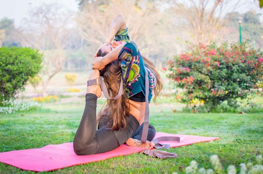 A woman is performing a yoga pose on a pink mat in a vibrant green garden. The pose involves bending backwards, with one leg stretched behind her head and both hands clasping her foot. She is wearing a colorful printed top and black leggings. The background features greenery, flowers, and soft sunlight filtering through the trees.