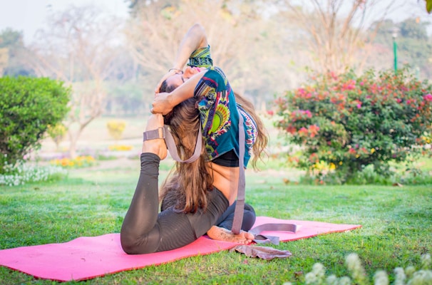 A woman is performing a yoga pose on a pink mat in a vibrant green garden. The pose involves bending backwards, with one leg stretched behind her head and both hands clasping her foot. She is wearing a colorful printed top and black leggings. The background features greenery, flowers, and soft sunlight filtering through the trees.