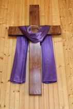 A wooden cross is affixed to a wooden wall, adorned with a purple cloth draped across the arms. A crown of thorns hangs at the top of the cross, symbolizing Christian imagery.