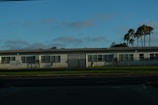 A single-story white building, possibly a preschool, with multiple windows is set against a backdrop of palm trees and a clear blue sky. The structure is surrounded by a low metal fence and is located next to a well-maintained grass lawn and a paved street in the foreground.