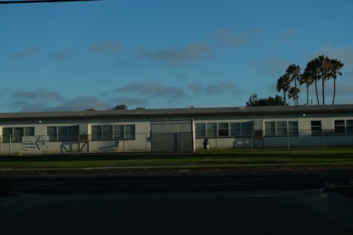 A single-story white building, possibly a preschool, with multiple windows is set against a backdrop of palm trees and a clear blue sky. The structure is surrounded by a low metal fence and is located next to a well-maintained grass lawn and a paved street in the foreground.