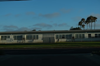 A single-story white building, possibly a preschool, with multiple windows is set against a backdrop of palm trees and a clear blue sky. The structure is surrounded by a low metal fence and is located next to a well-maintained grass lawn and a paved street in the foreground.