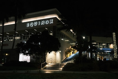 A modern building at night with illuminated branding for a business named Equinox. The structure features large windows and a staircase with bright lights lining each step. Palm trees partially obscure the view, and decorative lights wrap around a tree providing a warm ambiance.