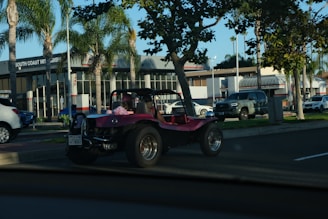A pink dune buggy is parked on the side of a road near a car dealership with palm trees lining the street. The dealership has a modern design with large windows. Several other vehicles are also visible, including a silver pickup truck and a white SUV. The setting appears to be a sunny day.