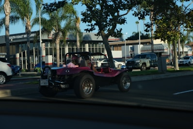 A pink dune buggy is parked on the side of a road near a car dealership with palm trees lining the street. The dealership has a modern design with large windows. Several other vehicles are also visible, including a silver pickup truck and a white SUV. The setting appears to be a sunny day.