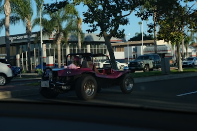 A pink dune buggy is parked on the side of a road near a car dealership with palm trees lining the street. The dealership has a modern design with large windows. Several other vehicles are also visible, including a silver pickup truck and a white SUV. The setting appears to be a sunny day.