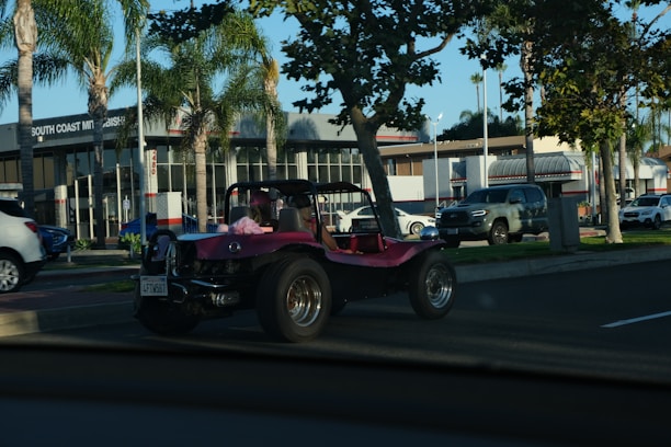 A pink dune buggy is parked on the side of a road near a car dealership with palm trees lining the street. The dealership has a modern design with large windows. Several other vehicles are also visible, including a silver pickup truck and a white SUV. The setting appears to be a sunny day.