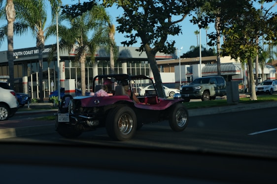 A pink dune buggy is parked on the side of a road near a car dealership with palm trees lining the street. The dealership has a modern design with large windows. Several other vehicles are also visible, including a silver pickup truck and a white SUV. The setting appears to be a sunny day.