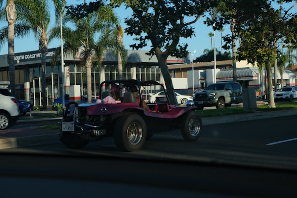 A pink dune buggy is parked on the side of a road near a car dealership with palm trees lining the street. The dealership has a modern design with large windows. Several other vehicles are also visible, including a silver pickup truck and a white SUV. The setting appears to be a sunny day.