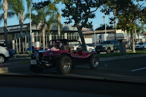 A pink dune buggy is parked on the side of a road near a car dealership with palm trees lining the street. The dealership has a modern design with large windows. Several other vehicles are also visible, including a silver pickup truck and a white SUV. The setting appears to be a sunny day.