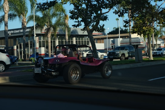 A pink dune buggy is parked on the side of a road near a car dealership with palm trees lining the street. The dealership has a modern design with large windows. Several other vehicles are also visible, including a silver pickup truck and a white SUV. The setting appears to be a sunny day.