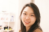 A young woman with long dark hair is smiling warmly at the camera. She is indoors, standing close to white shelves filled with miscellaneous items, including a blue spray bottle and some jars.