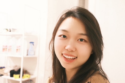 A young woman with long dark hair is smiling warmly at the camera. She is indoors, standing close to white shelves filled with miscellaneous items, including a blue spray bottle and some jars.