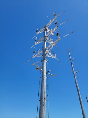 A tall, industrial electric utility pole against a clear blue sky. The pole is equipped with multiple cross arms and insulators, supporting electrical wires. It is one of a series of similar poles extending into the distance.