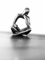 A minimalist sculpture of a human figure deeply engrossed in reading a book. The figure is crafted from a smooth, metallic material and adopts a seated pose with one hand resting under the chin, embodying a contemplative mood. The background is a soft gradient of black and white, enhancing the sculpture's reflective surface.