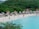 A scenic beach with white sand, lined with several thatched-roof umbrellas and lounge chairs. People are enjoying various activities like swimming and walking along the shore. Dense green foliage and hills form the backdrop, enhancing the tropical atmosphere.