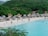A scenic beach with white sand, lined with several thatched-roof umbrellas and lounge chairs. People are enjoying various activities like swimming and walking along the shore. Dense green foliage and hills form the backdrop, enhancing the tropical atmosphere.