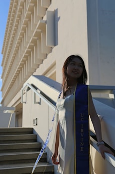 A person in a white dress and a blue and gold sash stands on a staircase, with a large building featuring geometric architectural designs in the background. The sash includes the text 'UC Irvine'. The image is taken from a side angle, showing shadows and sunlight creating a dynamic effect.