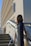 A person in a white dress and a blue and gold sash stands on a staircase, with a large building featuring geometric architectural designs in the background. The sash includes the text 'UC Irvine'. The image is taken from a side angle, showing shadows and sunlight creating a dynamic effect.