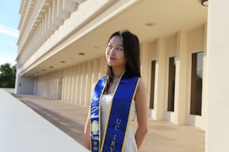 A young woman in a white dress stands outdoors, wearing a blue and yellow graduation stole with 'UC Irvine' and 'Class of 2023' printed on it. The backdrop features a modern concrete building with regularly spaced columns and rectangular windows. The woman gazes slightly to her right under a mostly clear sky.
