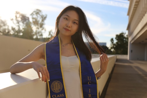 A young woman is wearing a graduation stole with the letters 'U C I R'. She is standing outdoors, leaning against a white wall. The setting sun casts a warm glow, and there are trees in the background.