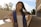 A young woman is wearing a graduation stole with the letters 'U C I R'. She is standing outdoors, leaning against a white wall. The setting sun casts a warm glow, and there are trees in the background.