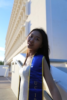 A young person wearing a graduation sash stands near a modern building with a repetitive architectural pattern. The sunlight creates dramatic shadows, emphasizing the bright blue and gold colors of the sash.