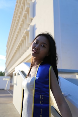 A young person wearing a graduation sash stands near a modern building with a repetitive architectural pattern. The sunlight creates dramatic shadows, emphasizing the bright blue and gold colors of the sash.