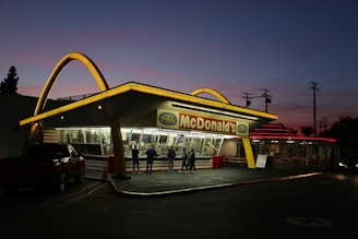 An iconic fast-food restaurant with bright yellow neon arches on the roof, set against a twilight sky. The building has a classic retro design with large glass windows and a red and yellow color scheme. Several people are gathered near the entrance, and a parked vehicle is visible in the foreground.