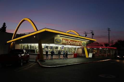 An iconic fast-food restaurant with bright yellow neon arches on the roof, set against a twilight sky. The building has a classic retro design with large glass windows and a red and yellow color scheme. Several people are gathered near the entrance, and a parked vehicle is visible in the foreground.