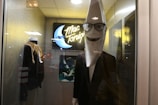 A mannequin with a large crescent moon head wearing sunglasses and a suit stands in a display case. There is a neon sign in the background with the text 'Mac Tonight' alongside a picture of a crescent moon playing a piano. A jacket on a mannequin is also visible beside it.