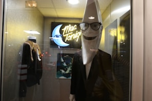 A mannequin with a large crescent moon head wearing sunglasses and a suit stands in a display case. There is a neon sign in the background with the text 'Mac Tonight' alongside a picture of a crescent moon playing a piano. A jacket on a mannequin is also visible beside it.