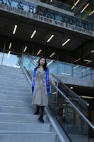 A person wearing a long beige coat and a blue and yellow graduation stole is walking down a flight of concrete stairs. The background features the facade of a modern building with large glass windows and signage indicating it is a science and engineering school.