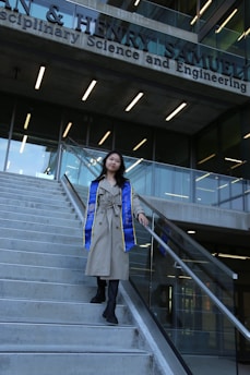 A person wearing a long beige coat and a blue and yellow graduation stole is walking down a flight of concrete stairs. The background features the facade of a modern building with large glass windows and signage indicating it is a science and engineering school.