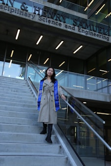 A person wearing a long beige coat and a blue and yellow graduation stole is walking down a flight of concrete stairs. The background features the facade of a modern building with large glass windows and signage indicating it is a science and engineering school.