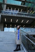 A person stands on a set of concrete stairs in front of a modern building with a clear glass facade. The building has a sign with the words 'Interdisciplinary Science and Engineering'. The individual is wearing a long beige coat and a blue sash that reads 'UC IRVINE'. They pose confidently, looking upwards.