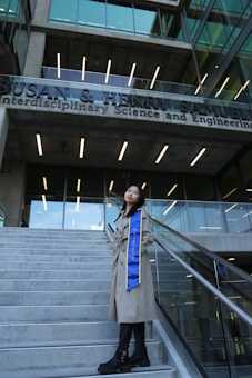 A person stands on a set of concrete stairs in front of a modern building with a clear glass facade. The building has a sign with the words 'Interdisciplinary Science and Engineering'. The individual is wearing a long beige coat and a blue sash that reads 'UC IRVINE'. They pose confidently, looking upwards.